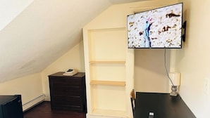 A room with a sloped ceiling, a television mounted on the wall, a wooden table, a chair, a small cabinet, and a heater on the floor.