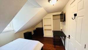 A small, compact room with a bed, a desk, and a chair. There is a door with a handle and a light fixture on the ceiling.