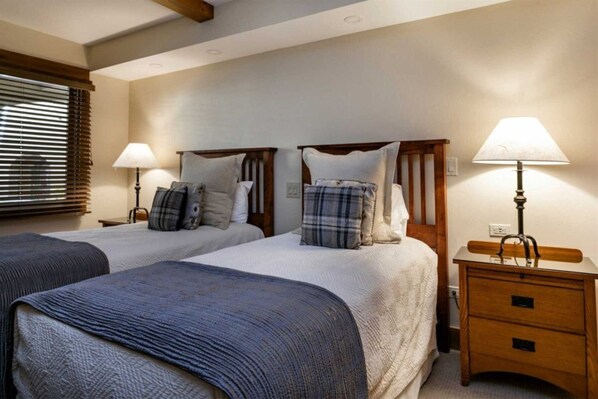 A hotel room with two single beds, wooden headboards, bedside tables with lamps, and a window with blinds.