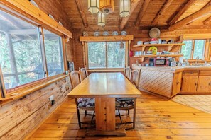 A wooden cabin interior with a large dining table, wooden chairs, a kitchen area with a microwave, and a window overlooking a forest.