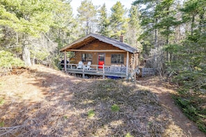 A rustic cabin with a wooden deck, two chairs, and a red door.