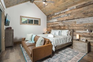 Une chambre avec un plafond en pente, un lit recouvert d’une couvre-lit à motifs, un canapé en cuir marron, une table de chevet avec une lampe et un mur en bois.
