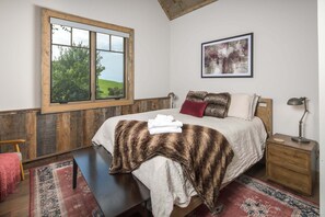 Quarto com uma grande janela, cama com manta de pele, mesa de cabeceira com abajur e tapete.