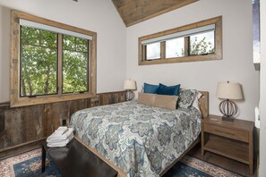 Ein Schlafzimmer mit einem großen Bett, zwei Nachttischen, einer Bank und einem Fenster mit Blick auf Bäume.