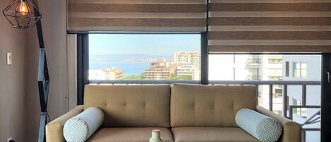 Sala de estar moderna com sofá bege, mesa de centro e vista para o mar e edifícios.