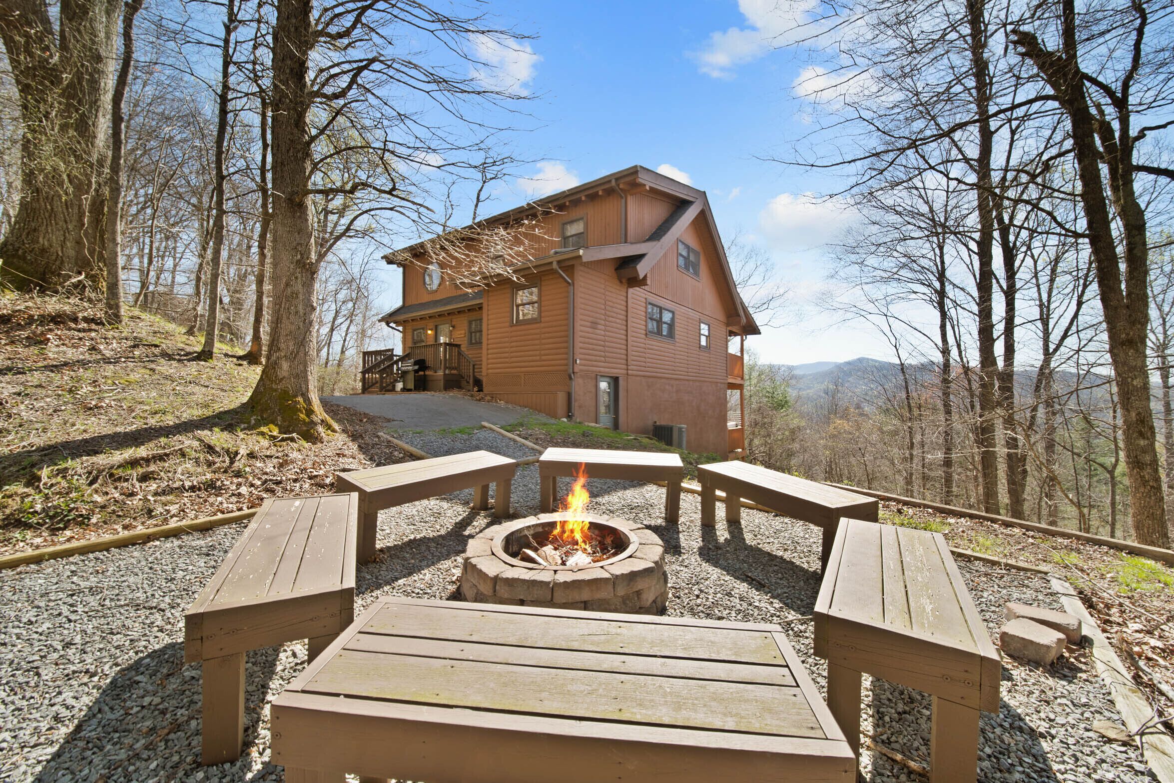 Eine Hütte mit Feuerstelle, umgeben von hölzernen Bänken, in einer bewaldeten Gegend mit Blick auf ein Gebirge.