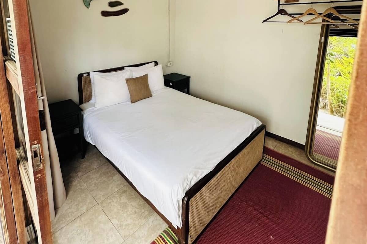 A bedroom with a bed, a ceiling fan, a nightstand, a wall-mounted shelf, and a window with a view of greenery.