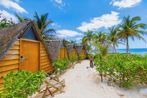 Een rij houten strandhutten met rieten daken, een zandpad en palmbomen.