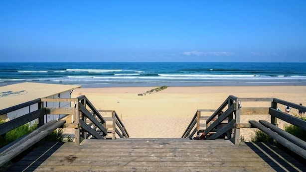 Ein hölzerner Steg führt zu einem Sandstrand, im Hintergrund sind Meereswellen zu sehen.