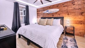 A modern bedroom with a wooden accent wall, a ceiling fan, a bed with white bedding, a small bedside table with a lamp, and a surfboard decoration.