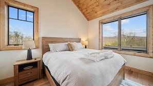 A bedroom with a wooden ceiling, a large bed with white bedding, two bedside tables with lamps, and large windows offering a view of the outdoors.