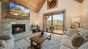 A cozy living room with a stone fireplace, a flat-screen TV mounted on the wall, a wooden ceiling, and a large sliding glass door leading to an outdoor deck.