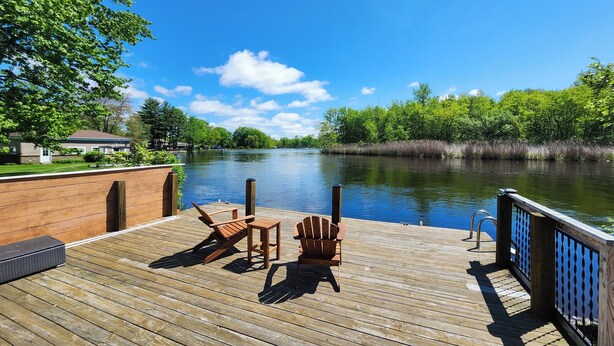 A wooden deck with a view of a river, surrounded by greenery and a house in the background.