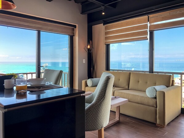 A modern room with a large window overlooking the sea, a sofa, a chair, and a small table with breakfast items.