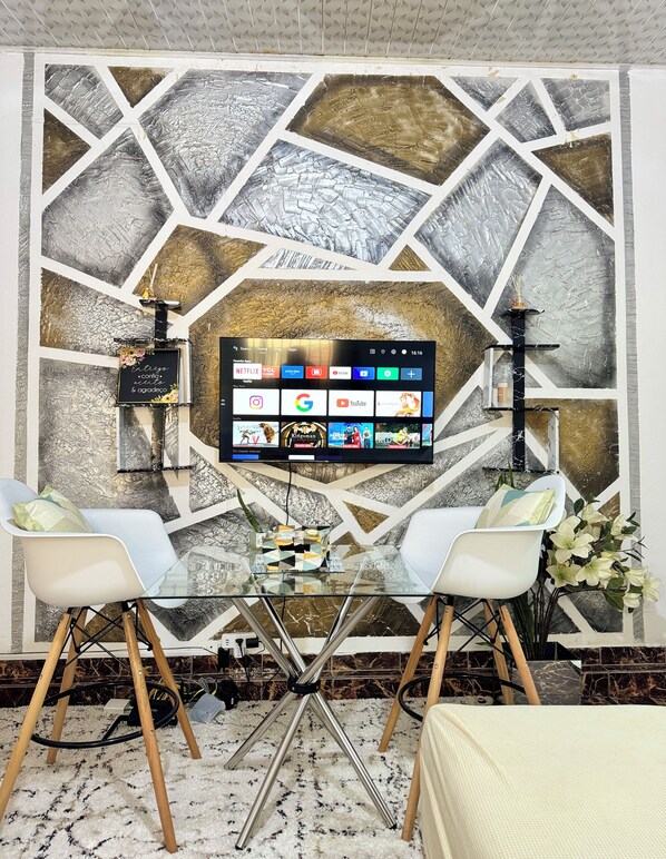 A modern living room with a large flat-screen TV mounted on a geometric-patterned wall, two white chairs, a glass coffee table, and a small decorative plant.