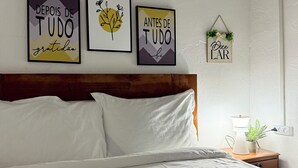 A neatly made bed with white linens in a room with a wooden headboard. Three framed artworks are hung on the wall above the bed, and a small potted plant is on the bedside table. - Charming 1BR Apt in the Heart of Maculusso-Free Netflix-High speed wifi (Luanda)