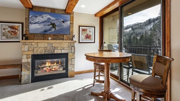 A cozy room with a stone fireplace, a round wooden table, and chairs. There's a large window offering a view of a snowy mountain. - Centrally Located 3 BR in Lionshead Village w/ Pool, Walk to Lift! (Vail)