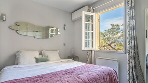 A bedroom with a bed, a window with curtains, a wall-mounted mirror, and a radiator. - Le Clos Sainte Anne by Interhome (Saint-Tropez)