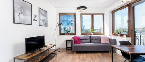 A modern living room with a flat-screen TV, a grey sofa, a wooden coffee table, and a dining table with chairs. There are two large windows with views of a cityscape.