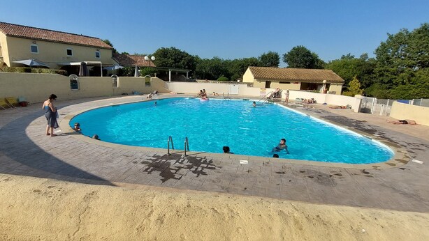 Une piscine avec une terrasse carrelée, entourée d’un petit muret et bordée de quelques bâtiments en arrière-plan.