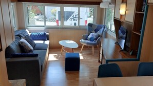 Un salon confortable avec un canapé, une petite table ronde et un fauteuil. On y trouve également un téléviseur posé sur un meuble et une fenêtre donnant sur l’extérieur.