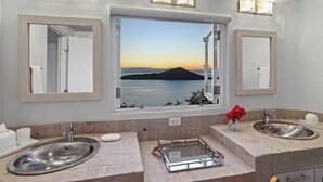 Ein Badezimmer mit zwei Waschbecken, einem großen Spiegel und einem Fenster mit einem malerischen Sonnenuntergang über dem Wasser.