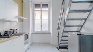 Eine schmale Küche mit weißen Schränken, einem Fenster, einem Edelstahlkühlschrank und einer Metallleiter.