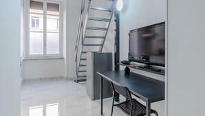 Ein kleines Zimmer mit Fernseher, Schreibtisch, Stuhl, einer Klapptreppe und einem Kühlschrank.