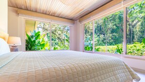 A bedroom with a large bed, a nightstand, and a window with a view of greenery.