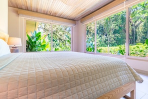 A bedroom with a large bed, a nightstand, and a window with a view of greenery.