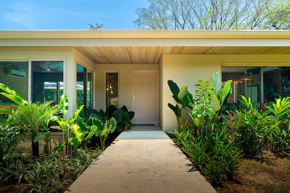 A modern house with a concrete pathway leading to a door, surrounded by lush tropical plants. - Chalet Tropical by Pura Villas (Uvita)