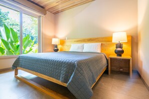 A bed with a wooden frame and a quilted bedspread, flanked by two bedside tables with lamps, in a room with a large window and a view of greenery.
