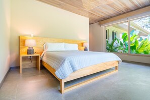 A modern bedroom with a wooden bed, bedside lamps, and a view of lush greenery through large windows. - Chalet Tropical by Pura Villas (Uvita)