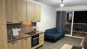 Una cocina moderna con armarios de madera, horno empotrado y lavamanos. Hay un sofá azul y una mesita con una lámpara. Se ve un balcón con barandilla y vistas al exterior.