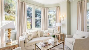 Un salon bien éclairé, avec un canapé, des fauteuils et une table basse. De grandes fenêtres, dotées de rideaux, laissent entrer la lumière naturelle et offrent une vue sur la verdure environnante.