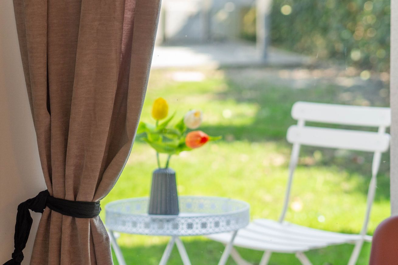 Une fenêtre avec un rideau retenu, une petite table sur laquelle est posé un vase de fleurs et une chaise blanche, le tout donnant sur un jardin.
