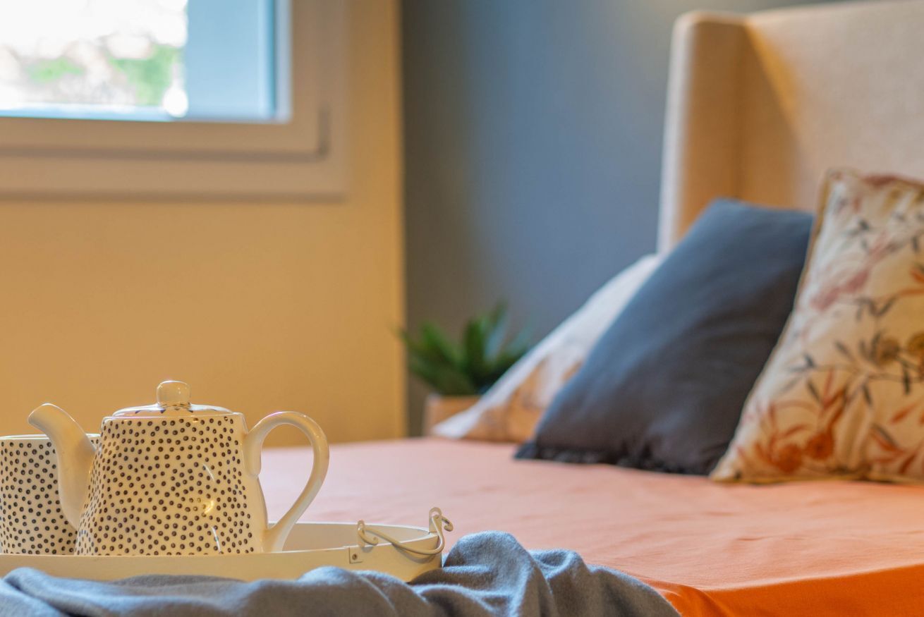 A bed with a gray blanket, a white tray with teapots, and a potted plant in a room with a window.