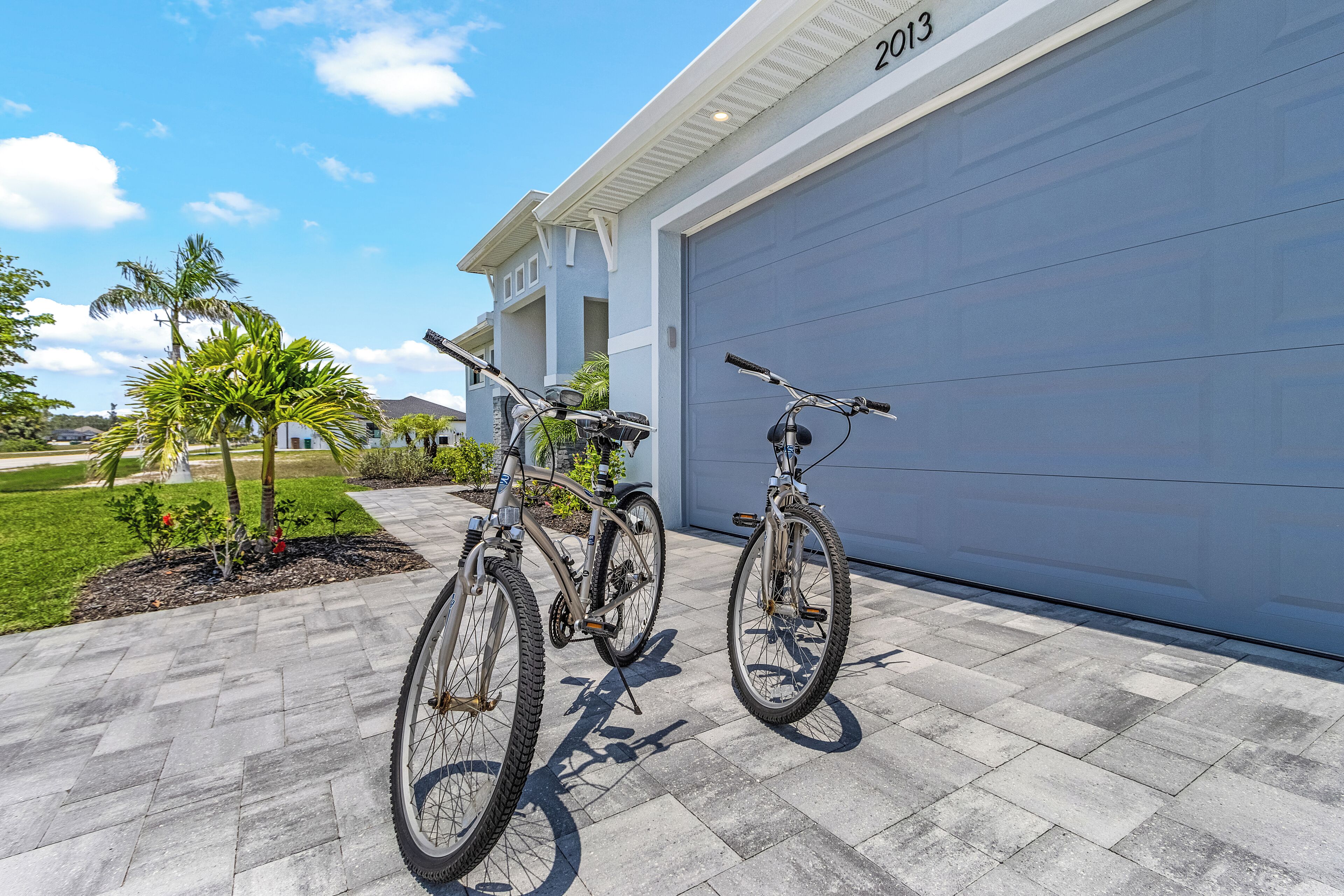 Een modern huis met een garage deur waarop 2013 staat, twee fietsen voor de deur en een verzorgd gazon met palmboompjes.