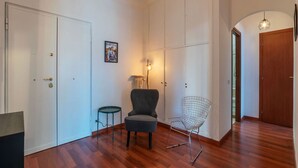 A room with wooden flooring, a grey armchair, a wireframe chair, a small round table, and a wall-mounted lamp.