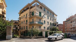 A multi-story residential building with balconies, a car parked in front, and a clear sky.
