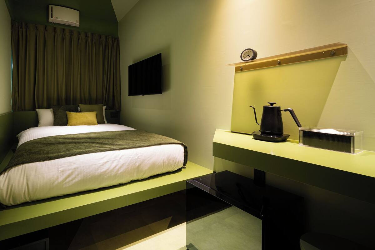 A modern hotel room with a bed, a bedside table with a coffee maker, a flat-screen TV, and a window with curtains.