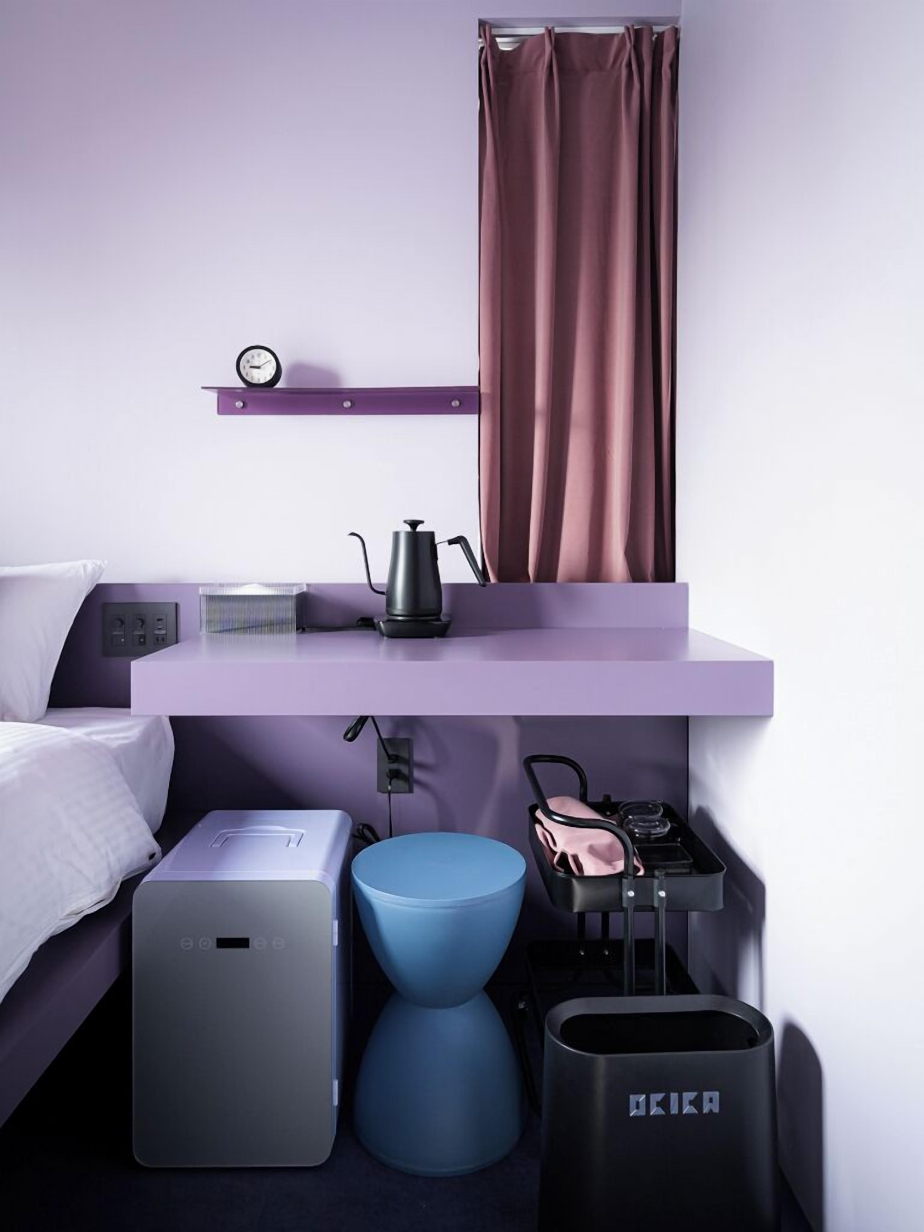 A modern hotel room with a bed, a small desk with a kettle, a trash bin, and a compact kitchenette.