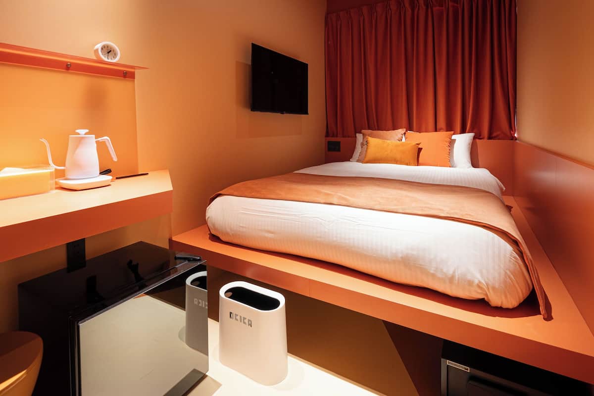 A hotel room with a bed, a desk, a chair, a TV, and a trash bin.