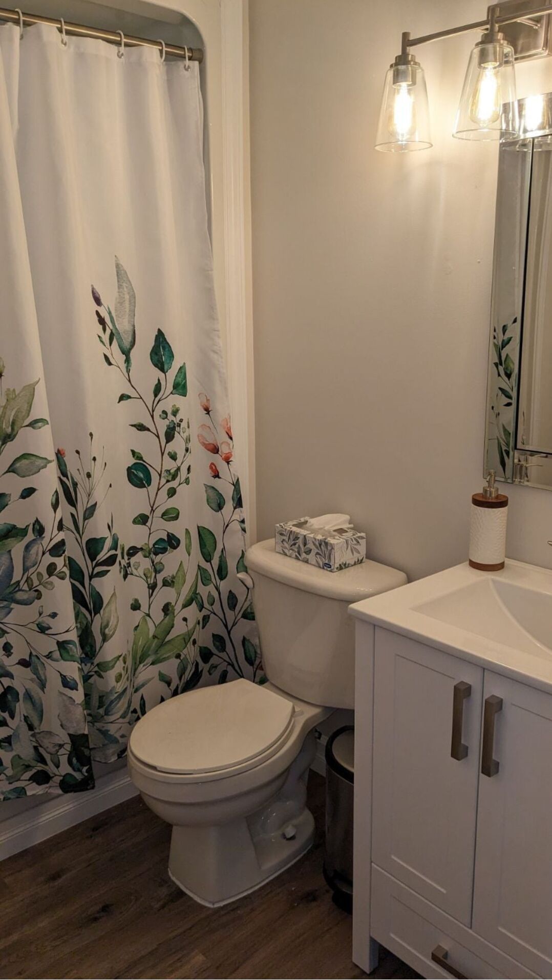 A bathroom with a toilet, a shower curtain with floral patterns, a toilet paper holder, and a cabinet with drawers.