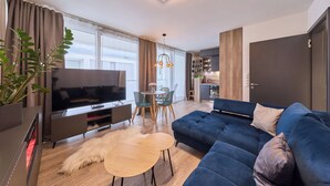 Una sala de estar moderna con un televisor de pantalla plana grande, un sofá azul, una pequeña mesa redonda y un comedor con sillas.