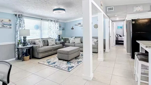 A spacious living area with a sofa, ottoman, and a dining table with chairs. There is a kitchen area visible in the background.