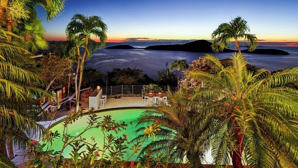 A pool area with a view of the ocean at sunset, surrounded by palm trees and a deck with seating.