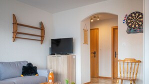 A living room with a sofa, a coffee table, a TV, a dartboard, and a wooden chair. - Austral 105 by Interhome (Ovronnaz)