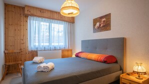 A bedroom with a bed, a wooden nightstand with a lamp, a window with sheer curtains, and a wall-mounted picture. - Austral 105 by Interhome (Ovronnaz)