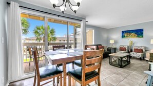 A spacious living area with a dining table, chairs, and a sofa. There is a large window with a view of palm trees and a building. - Sandpiper Cover 2031 near beach sleeps 4 (Destin)